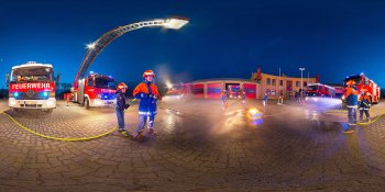 Nighttime firefighting scene with a tall ladder truck raised and firefighters coordinating on a cobblestone square.