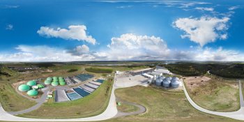 Aerial view of an industrial facility with green-roofed tanks, silver silos, solar panels, and roads across a grassy landscape under a blue sky.