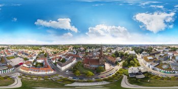 Panoramic aerial view of a small town with red-roof houses, curved streets, and a central church tower under a blue sky.