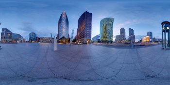 Panoramic view of a modern city skyline along a waterfront promenade at dusk, with illuminated glass skyscrapers reflecting on the water.
