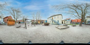 Panoramic view of a sandy public square with leafless trees, playground equipment, and a curved white building in the background.